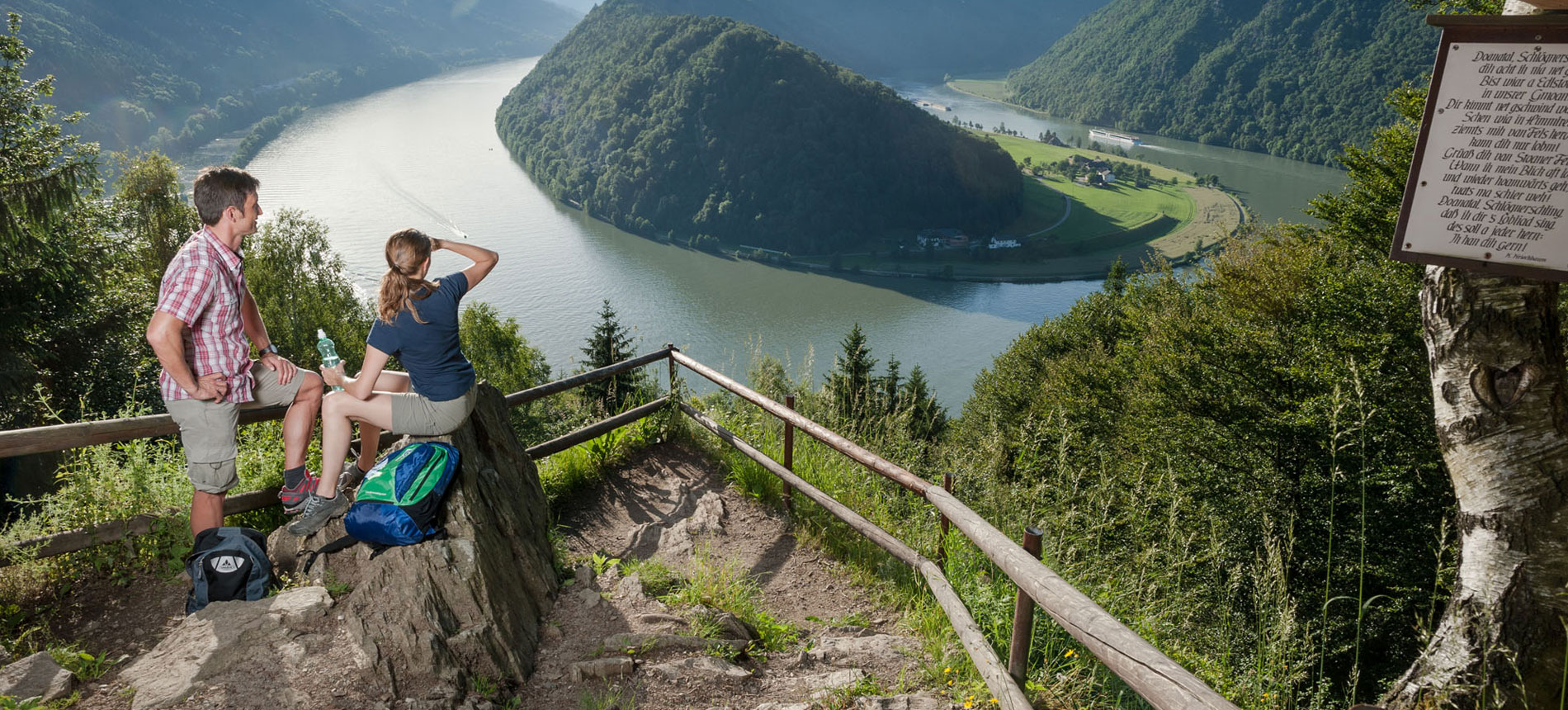 La fotografia mostra due turisti che, durante una passeggiata in montagna, si fermano ad ammirare, dall'alto, l'ansa di un fiume.